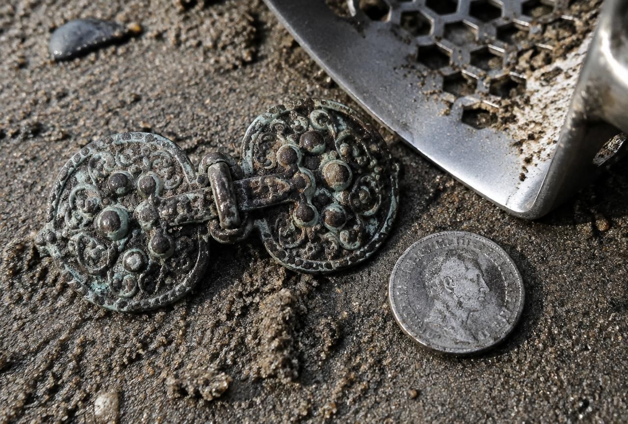 Moneda de plata oxidada y broche de plata labrado sobre arena húmeda de playa, junto a una cuchara de excavación